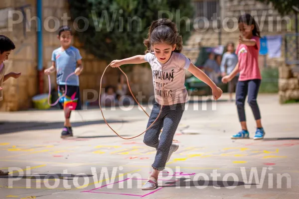 ילדים מדלגים בחבל ומשחקים בחבל בחצר בית ספר ממדינת ישראל. צַעֲצוּעַ, מִכְנָסַיִים קְצָרִים, עֵץ